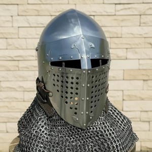 May include: A silver-colored medieval helmet with a riveted visor and a chainmail neck guard. The helmet has a rounded top and a series of small holes for ventilation. The chainmail is draped over a wooden surface.