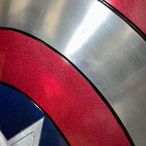 May include: Close-up of a Captain America shield. The shield features a metallic silver center, bordered by a red ring with a glittery texture. A blue section with a white star is visible.