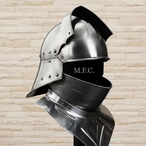 May include: A polished, silver-colored medieval helmet with a visor and neck guard. The helmet has rivets and a curved top, with the text "M.F.C." visible on the front. The design suggests historical armor.