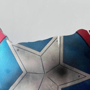 May include: Fragment of a shield with a star design. The shield features a blue center with a silver star, and red and blue edges. The shield has a distressed appearance, with visible wear and tear.