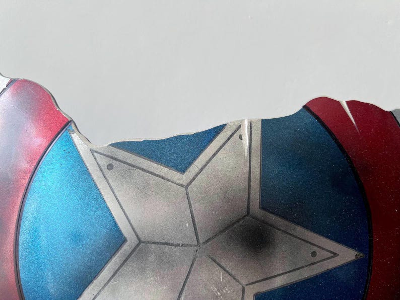May include: Fragment of a shield with a star design. The shield features a blue center with a silver star, and red and blue edges. The shield has a distressed appearance, with visible wear and tear.