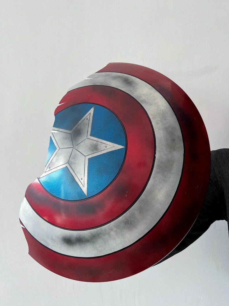 May include: A Captain America shield prop, featuring a red, white, and blue color scheme. The shield has a central blue circle with a white star, surrounded by red and white stripes. The shield is slightly weathered.