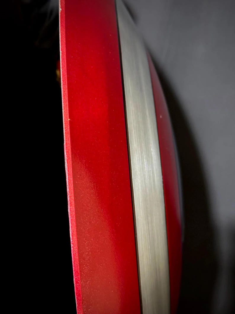 May include: A red and silver shield with a glossy finish. The shield has a bright red section and a silver stripe. The edges are smooth and the overall shape is rounded.