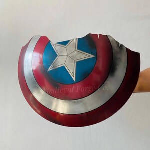 May include: A distressed, battle-worn shield with a red, white, and blue color scheme. The shield features a central blue circle with a white star, surrounded by concentric red and silver bands. The edge is jagged, and the text "Medieval Forge Co." is visible.