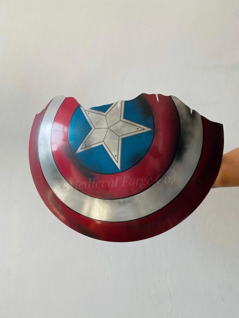 May include: A distressed, battle-worn shield with a red, white, and blue color scheme. The shield features a central blue circle with a white star, surrounded by concentric red and silver bands. The edge is jagged, and the text "Medieval Forge Co." is visible.