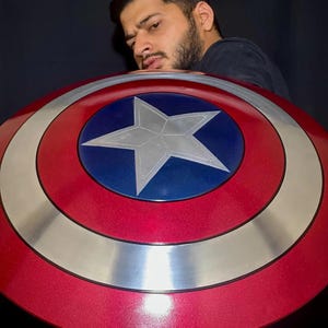 May include: A red, white, and blue Captain America shield with a star in the center. The shield has a metallic silver ring and a glossy finish. The shield is a prop or collectible item.