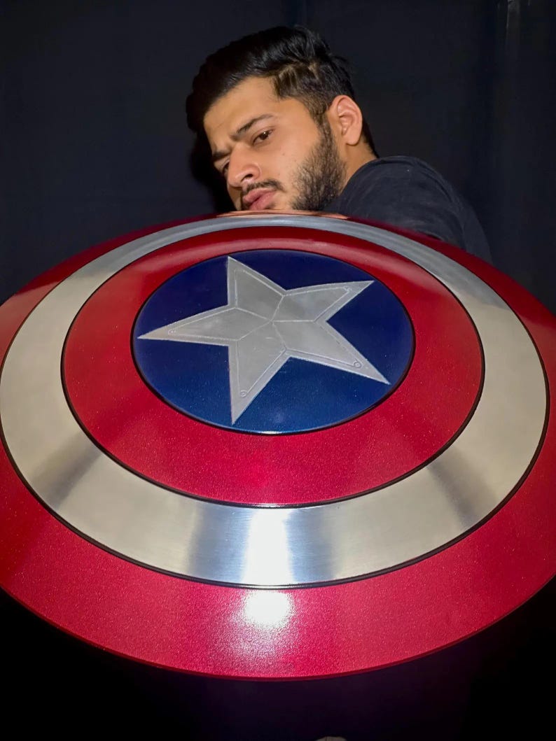 May include: A red, white, and blue Captain America shield with a star in the center. The shield has a metallic silver ring and a glossy finish. The shield is a prop or collectible item.