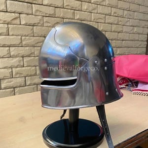 May include: A polished steel helmet with a rounded top and a visor. The helmet is riveted and has a chin strap. The helmet is displayed on a black stand. The text "medievalforgeco." is visible on the helmet.