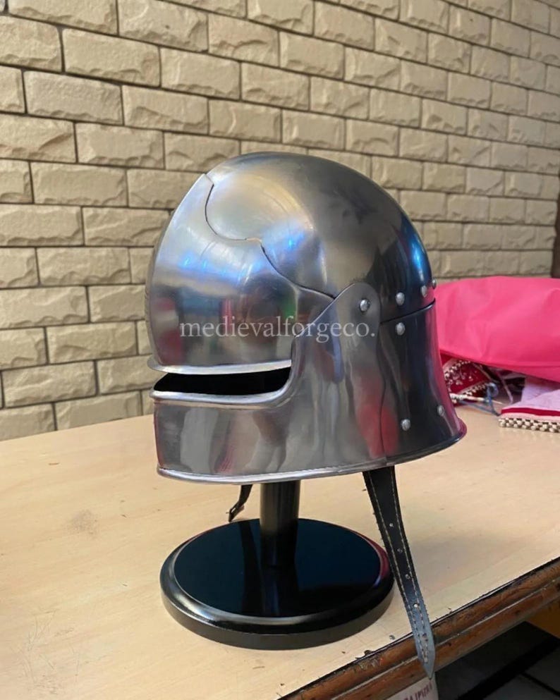 May include: A polished steel helmet with a rounded top and a visor. The helmet is riveted and has a chin strap. The helmet is displayed on a black stand. The text "medievalforgeco." is visible on the helmet.