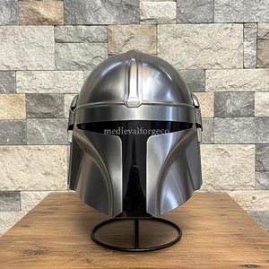 Op de afbeelding: Een zilverkleurige helm met een opvallend vizier en een afgeronde bovenkant. De helm wordt getoond op een zwarte standaard tegen een getextureerde stenen muur. De tekst "medievalforgeco" is zichtbaar op de helm.
