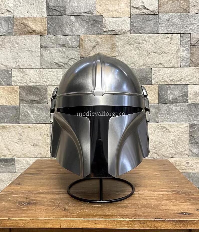 Op de afbeelding: Een zilverkleurige helm met een opvallend vizier en een afgeronde bovenkant. De helm wordt getoond op een zwarte standaard tegen een getextureerde stenen muur. De tekst "medievalforgeco" is zichtbaar op de helm.