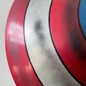 May include: Close-up of a Captain America shield. The shield features a red, white, and blue color scheme with a silver stripe and a white star on a blue background. The shield has a metallic finish.
