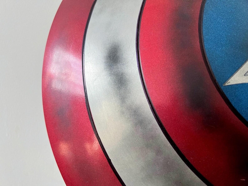 May include: Close-up of a Captain America shield. The shield features a red, white, and blue color scheme with a silver stripe and a white star on a blue background. The shield has a metallic finish.