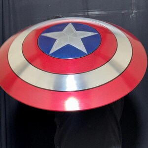 May include: A red, white, and blue shield with a silver star in the center. The shield has a circular design with concentric rings of red, white, and blue. The shield is a prop or costume accessory.