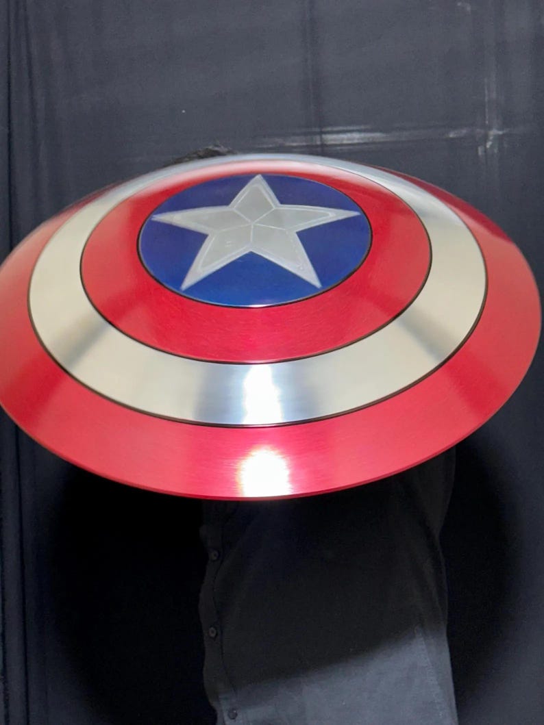 May include: A red, white, and blue shield with a silver star in the center. The shield has a circular design with concentric rings of red, white, and blue. The shield is a prop or costume accessory.