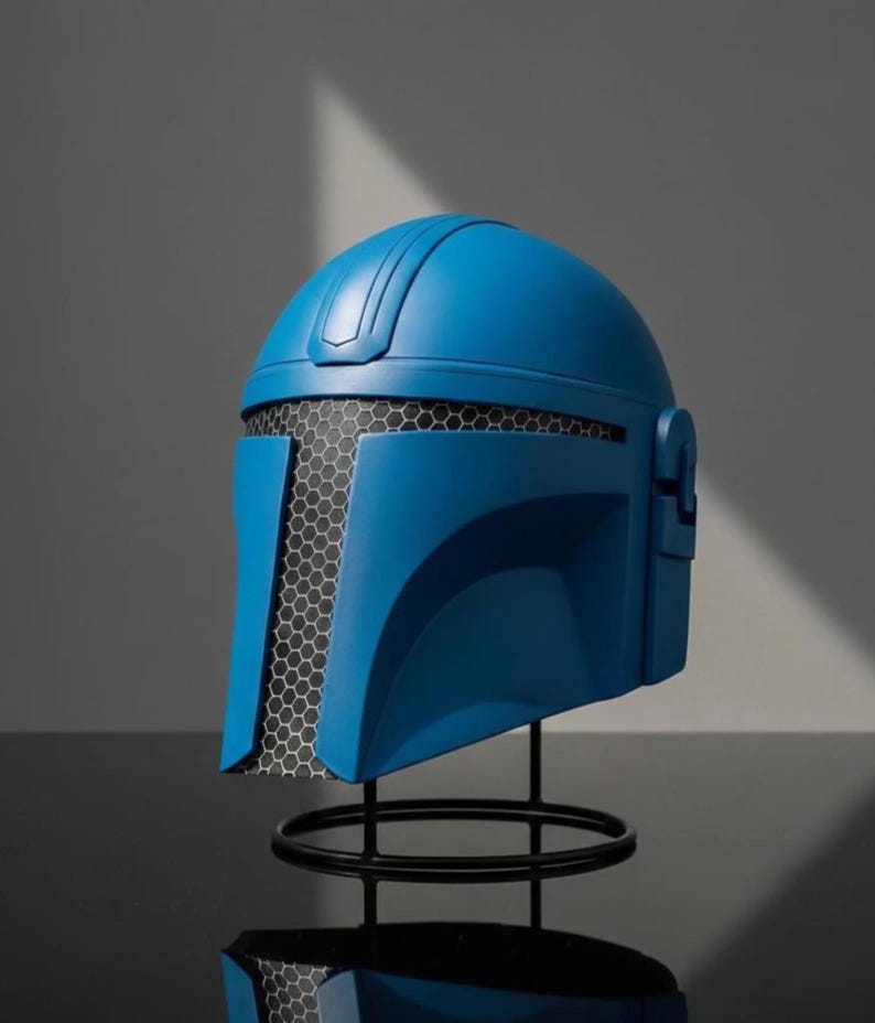 Puede incluir: Casco azul con visera de malla negra, exhibido sobre un soporte de metal negro. El casco tiene un acabado liso y mate y un dise&ntilde;o que recuerda a un guerrero futurista. El soporte es circular, proporcionando una base estable.