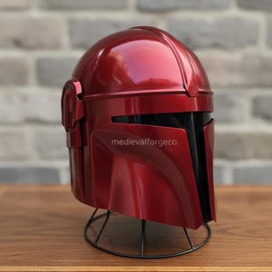 Op de afbeelding: Een metallic rode helm met een zwart vizier en oorbeschermers. De helm staat op een zwarte draadstandaard. De tekst "medievalforgeco." is zichtbaar. Waarschijnlijk een kostuum of verzamelobject.