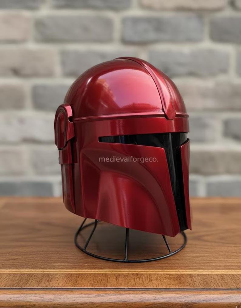 Op de afbeelding: Een metallic rode helm met een zwart vizier en oorbeschermers. De helm staat op een zwarte draadstandaard. De tekst "medievalforgeco." is zichtbaar. Waarschijnlijk een kostuum of verzamelobject.