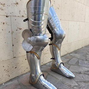 May include: A pair of polished steel leg armor, featuring articulated plates and decorative brass accents. The armor includes knee protection and pointed sabatons, designed for historical reenactment or display. The metal has a reflective surface.