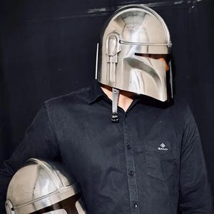 Może przedstawiać: Błyszczący, srebrny hełm Mandalorianina z wizjerem w kształcie litery T jest noszony. Drugi hełm trzymany jest w ramieniu osoby. Hełmy są metalowe i mają futurystyczny design. Osoba ma na sobie czarną koszulę z guzikami.