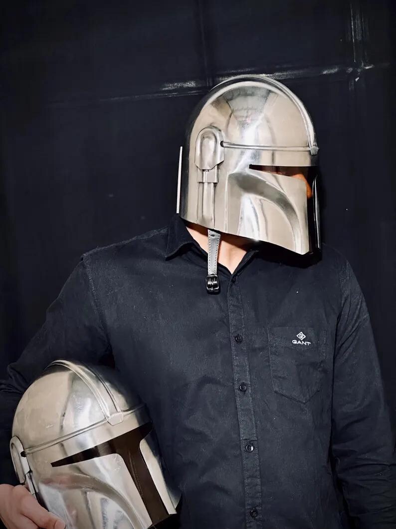 Może przedstawiać: Błyszczący, srebrny hełm Mandalorianina z wizjerem w kształcie litery T jest noszony. Drugi hełm trzymany jest w ramieniu osoby. Hełmy są metalowe i mają futurystyczny design. Osoba ma na sobie czarną koszulę z guzikami.