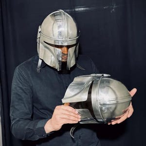 Może przedstawiać: Srebrny hełm Mandalorian i pasujący napierśnik. Hełm ma wizjer w kształcie litery T i zaokrągloną g&oacute;rę. Napierśnik ma owalny kształt z otwieraną g&oacute;rą. Osoba w czarnej koszuli trzyma przedmioty.