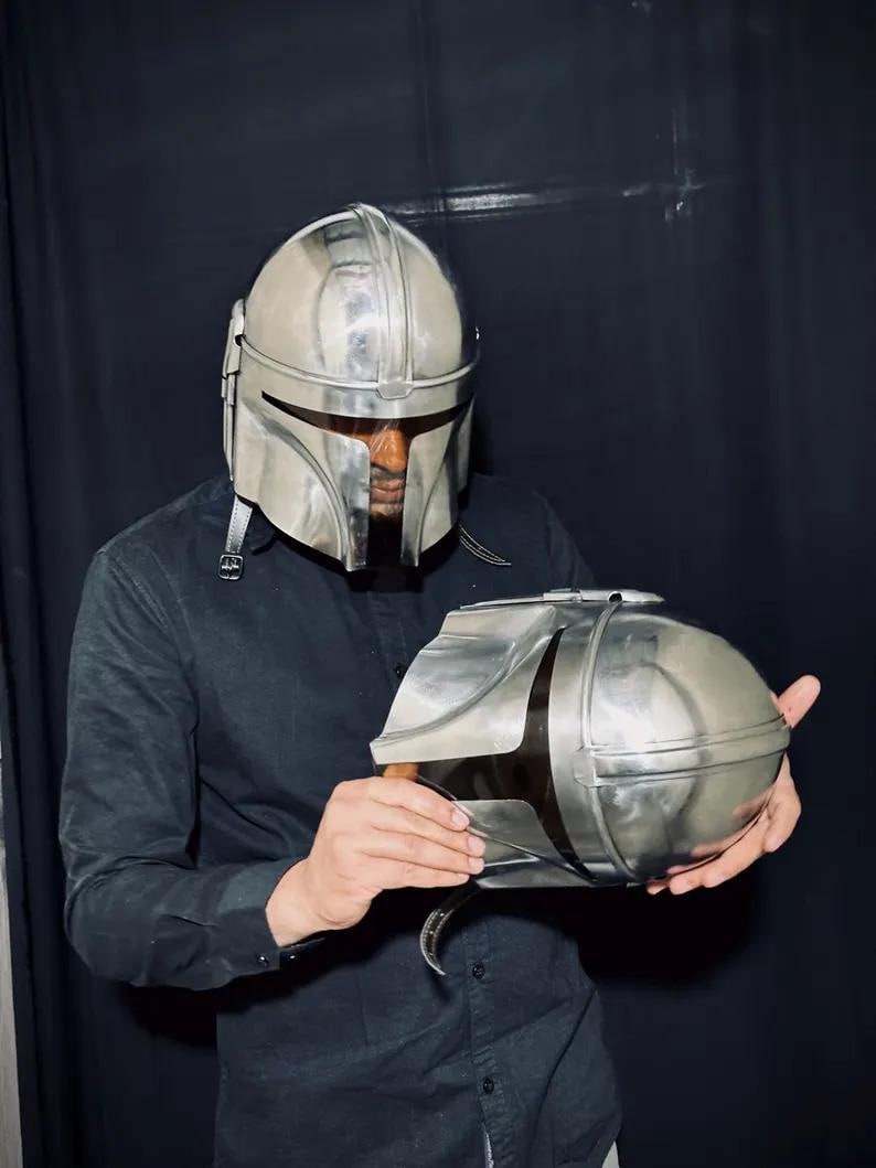 Może przedstawiać: Srebrny hełm Mandalorian i pasujący napierśnik. Hełm ma wizjer w kształcie litery T i zaokrągloną g&oacute;rę. Napierśnik ma owalny kształt z otwieraną g&oacute;rą. Osoba w czarnej koszuli trzyma przedmioty.