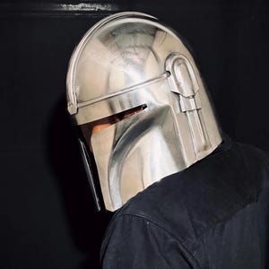 Może przedstawiać: Srebrny hełm Mandalorianina z wizjerem i kanciastym designem. Hełm wykonany jest z metalicznego materiału i odbija światło otoczenia. Jest częścią kostiumu lub cosplayu.