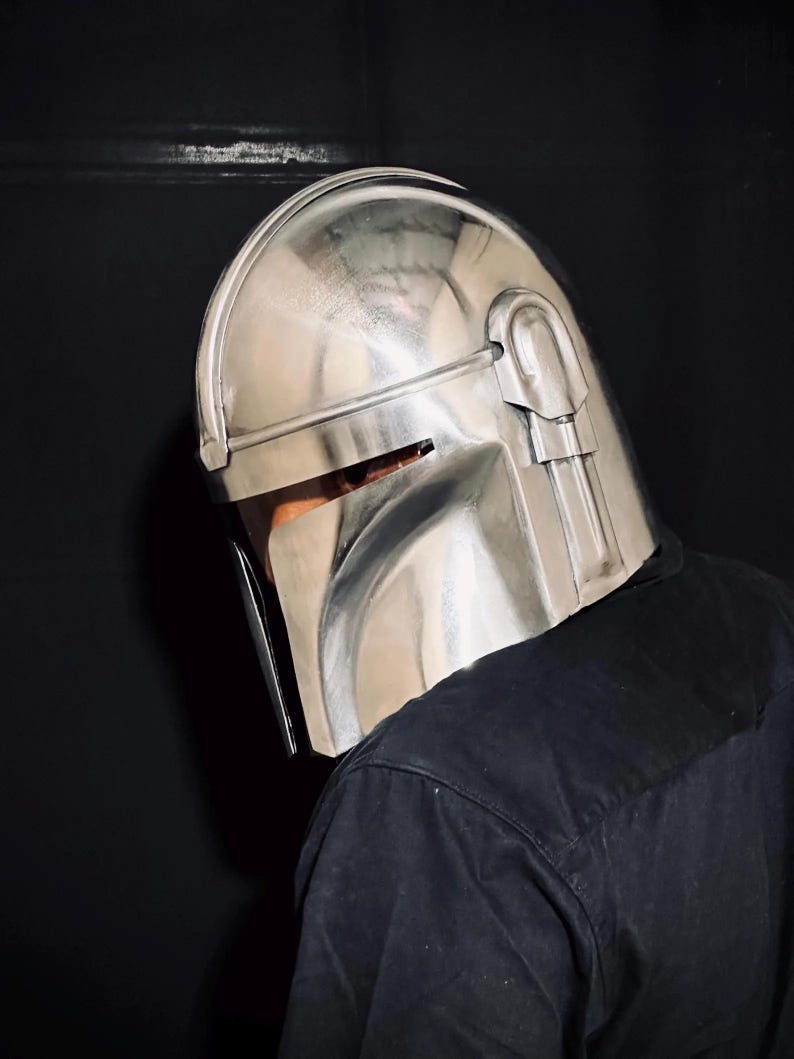 Może przedstawiać: Srebrny hełm Mandalorianina z wizjerem i kanciastym designem. Hełm wykonany jest z metalicznego materiału i odbija światło otoczenia. Jest częścią kostiumu lub cosplayu.