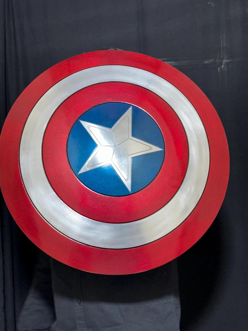 May include: A round shield with a red outer ring, a silver inner ring, and a blue center. A silver five-pointed star is in the center. The shield is a replica of Captain America's shield.