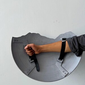 May include: A gray, semi-circular shield with a jagged top edge. The shield has two black straps with buckles for securing the arm. The shield is held by a hand.