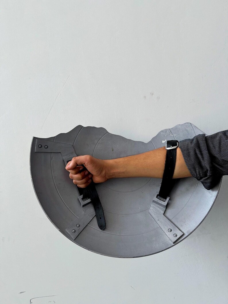 May include: A gray, semi-circular shield with a jagged top edge. The shield has two black straps with buckles for securing the arm. The shield is held by a hand.