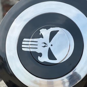 May include: A black and silver shield featuring a white skull emblem. The shield has a circular design with a silver border and a black center. The skull emblem is in the center of the shield.