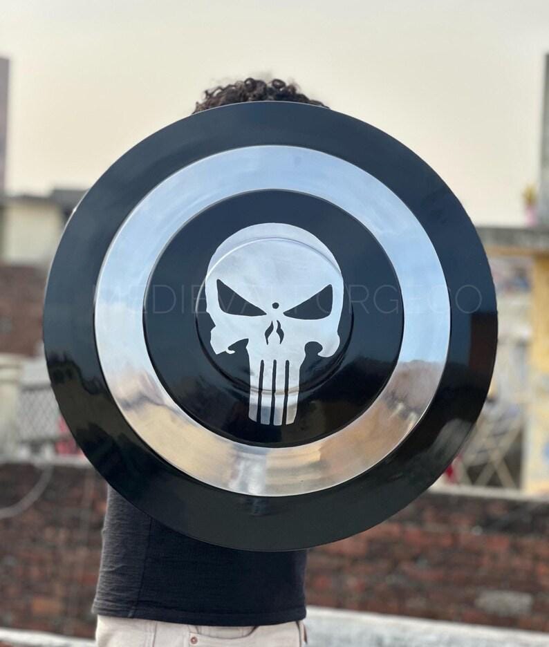 May include: A black and silver shield featuring a white skull emblem at the center. The shield has a glossy finish with concentric circles in black and silver. The shield is held up against a blurred outdoor background.