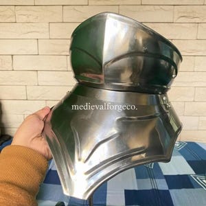 May include: A polished steel gorget, a piece of medieval armor, is held up. The gorget is made of multiple curved plates and is designed to protect the neck and upper chest. The text "medievalforgeco." is visible.