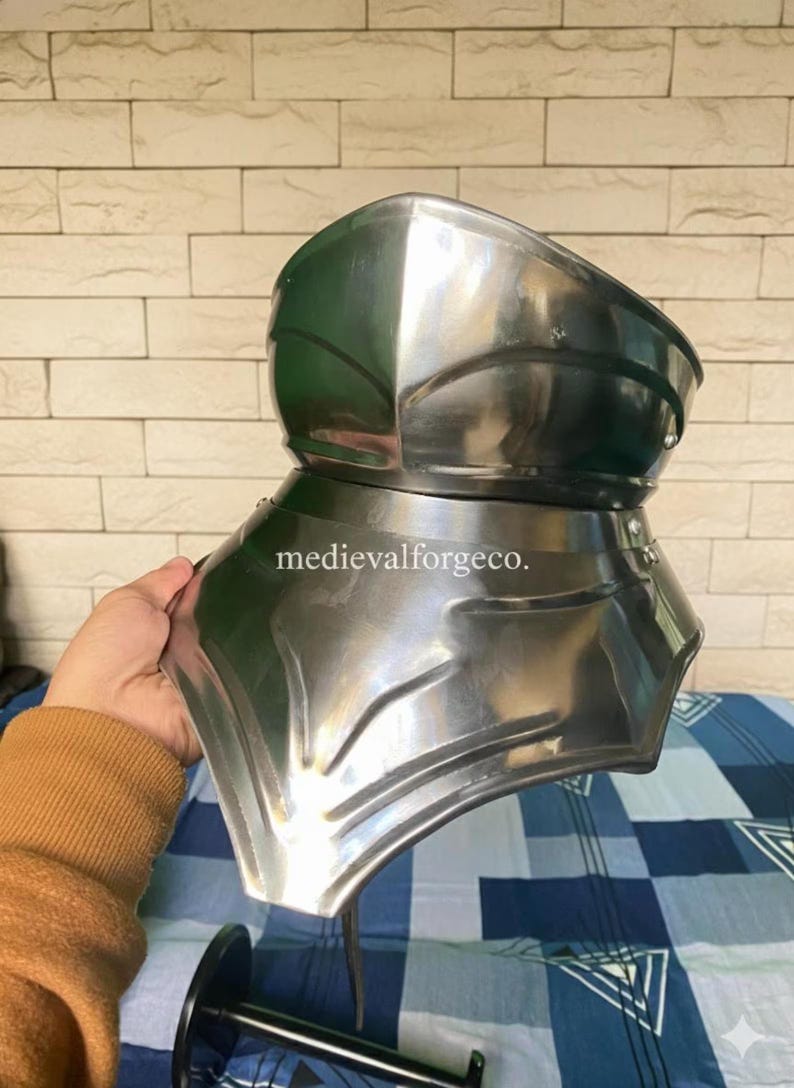May include: A polished steel gorget, a piece of medieval armor, is held up. The gorget is made of multiple curved plates and is designed to protect the neck and upper chest. The text "medievalforgeco." is visible.