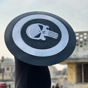 May include: A black and silver shield with a skull emblem. The shield has a circular design with a black outer ring and a silver inner circle. The skull emblem is white and centered on the shield.