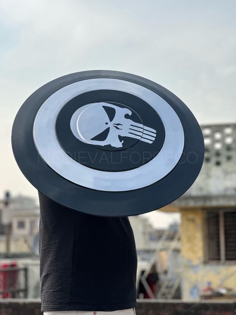 May include: A black and silver shield with a skull emblem. The shield has a circular design with a black outer ring and a silver inner circle. The skull emblem is white and centered on the shield.