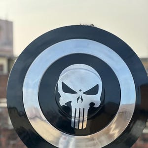 May include: A black and silver shield featuring a skull emblem. The shield has a circular design with a black center, a silver ring, and a black outer edge. The skull emblem is in the center.