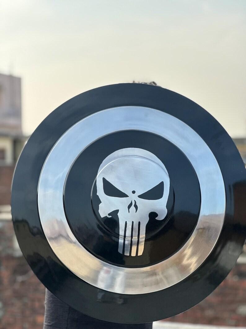 May include: A black and silver shield featuring a skull emblem. The shield has a circular design with a black center, a silver ring, and a black outer edge. The skull emblem is in the center.