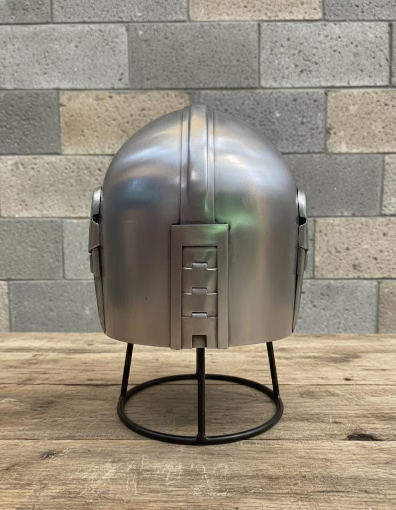 Op de afbeelding: Een zilverkleurige helm met een afgeronde bovenkant en rechthoekige details aan de achterkant. De helm wordt gepresenteerd op een zwarte metalen standaard met een cirkelvormige basis. De achtergrond toont een grijze bakstenen muur en een houten oppervlak.