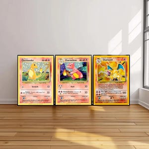 May include: Three framed Pokémon trading cards featuring Charmander, Charmeleon, and Charizard. The cards have colorful illustrations of the characters with their names and stats displayed. The frames are black, and the cards are displayed on a wooden floor.