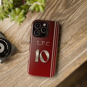 May include: A burgundy phone case with the letters "L.F.C" and the number "10" in silver. The case has vertical silver stripes on the sides. The phone case is on a wooden surface.
