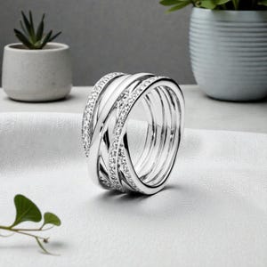 Puede incluir: Un anillo plateado con un diseño entrelazado. El anillo presenta múltiples bandas, algunas adornadas con pequeñas piedras brillantes. Se muestra sobre una superficie blanca, con un fondo borroso que incluye plantas en macetas.