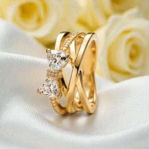 Può includere: Un anello color oro con un design a forma di fiocco. Il fiocco presenta due pietre preziose triangolari e trasparenti e piccole pietre di accento. L'anello ha un design a più bande ed è esposto su una superficie bianca e setosa.