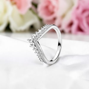 Può includere: Un anello color argento con un design a forma di V. L'anello è adornato con piccole pietre preziose scintillanti lungo i bordi, creando un aspetto a corona. L'anello è posto su uno sfondo bianco morbido con fiori rosa e bianchi sfocati.