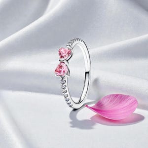 May include: A silver ring with two pink heart-shaped stones and a row of small clear stones. The ring is set against a white background with a pink petal.