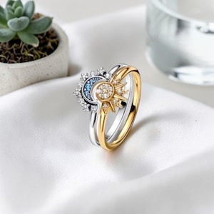 May include: A two-tone ring set featuring a celestial design. One ring is silver with a crescent moon and blue accents, the other is gold with a sunburst design. Both rings are adorned with small, sparkling stones.
