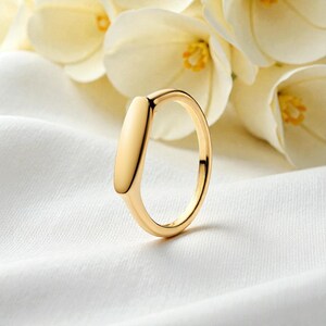 Puede incluir: Un anillo dorado con una parte superior rectangular, sobre un fondo de tela blanca. El anillo tiene un diseño minimalista con una superficie lisa y pulida. Suaves flores blancas en el fondo. El diseño del anillo es moderno.