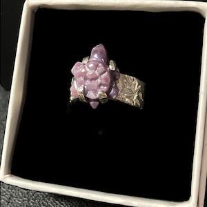 May include: A ring featuring a carved purple stone turtle centerpiece mounted on a textured silver-toned metal band. The turtle is secured by four metal prongs, and the band displays an embossed pattern across its surface, presented in a box.